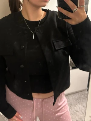Svart cropped cardiganjacka - Stilren svart cardiganjacka med rund halsringning och knäppning framtill. Jackan har två dekorativa fickor på bröstet och är i en rak, kortare modell med långa ärmar. Perfekt att slänga över en t-shirt för en clean look.