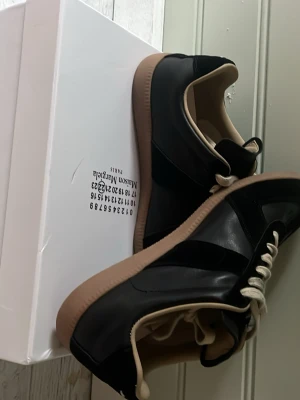 Svarta sneakers Maison Margiela - Stilrena svarta sneakers från Maison Margiela med beige sula och vita skosnören. Skorna har en kombination av mocka och läder, samt klassisk låg silhuett. Perfekta för dig som gillar minimalistisk och trendig design.