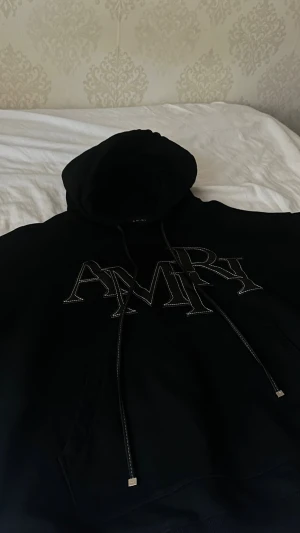 Svart hoodie med AMIRI-logga - Säljer en svart hoodie från AMIRI med stor vit logga framtill. Hoodien har huva med dragsko och en klassisk känguruficka. Perfekt för dig som gillar streetwear och vill ha en stilren men ändå statement look.