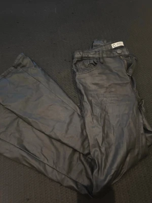 Svarta ledärbyxor - Säljer ett par svarta  ledärjeans med hög midja och vida ben. Jeansen har klassisk femficksmodell och knappgylf. Materialet har en glansig finish som ger en cool look. Perfekta för dig som vill sticka ut lite extra.