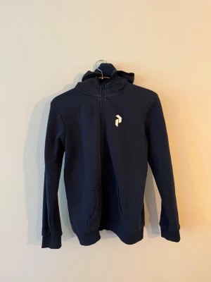 Mörkblå hoodie med vit logga Peak Performace - Snygg mörkblå hoodie med dragkedja och huva. Stor vit logga på bröstet och ryggen ger en clean och modern look. Hoodien har två fickor framtill och ribbade muddar vid ärmar och nederkant. Perfekt för chill dagar eller när du vill ha en enkel stil.