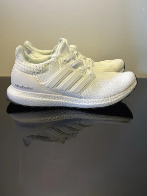 Vita Adidas Ultraboost sneakers - Säljer ett par helvita Adidas Ultraboost sneakers med stickad ovandel och klassiska tre ränder på sidan. Skorna har en bekväm Boost-sula och svart yttersula med Continental-gummi för extra grepp. Perfekta för dig som gillar stilrena och sportiga sneakers.