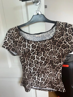 Leopardmönstrad topp med kort ärm - Säljer en snygg leopardmönstrad topp med korta ärmar och rund halsringning. Toppen har svarta dekorativa kanter vid halsen och är tillverkad i ett mjukt, stretchigt material. Perfekt för dig som vill sticka ut lite extra!