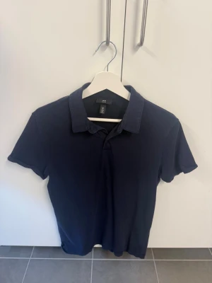 Mörkblå pikétröja från H&M - Klassisk mörkblå pikétröja från H&M i slim fit-modell. Tröjan har korta ärmar, krage och knappar vid halsen. Perfekt för en clean och enkel stil. Tillverkad i mjuk bomull för skön känsla.