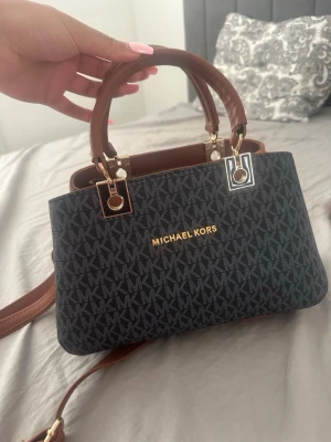Michael Kors monogram handväska - Snygg handväska från Michael Kors i mörkblått med brunt läder och guldiga detaljer. Väskan har MK-monogram över hela ytan, två handtag, justerbar axelrem och en praktisk ytterficka med dragkedja. Perfekt för dig som gillar klassisk och trendig design. Oanvänd 