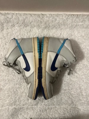 Nike Dunk High blå/vit sneakers - Nike Dunk High sneakers i vitt och ljusgrått läder med blå detaljer och swoosh i olika blå nyanser på varje sida. Skorna har högt skaft och klassisk gummisula med blå och turkosa inslag. Perfekta för dig som gillar retrostil och unika färgkombinationer.