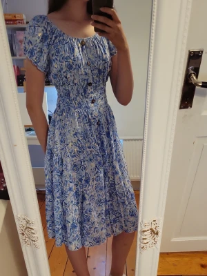 Blåvit blommig kortklänning XS - Söt blåvit klänning med blommigt mönster och korta puffärmar. Klänningen har smock i midjan, knappar framtill och en luftig kjol som slutar strax ovanför knäna. Perfekt för varma dagar och ger en romantisk känsla.