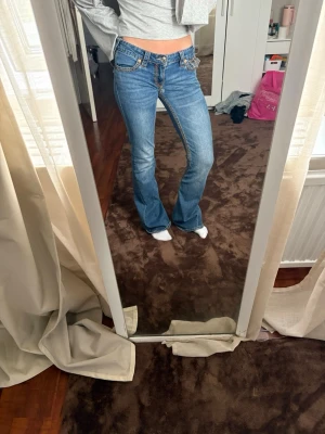 Blå bootcut jeans från True Religion - Säljer ett par blå bootcut jeans från True Religion med kontrastsömmar och detaljerade fickor bak. Jeansen har låg midja och klassisk femficksdesign. Perfekta för dig som gillar en avslappnad men ändå trendig stil.