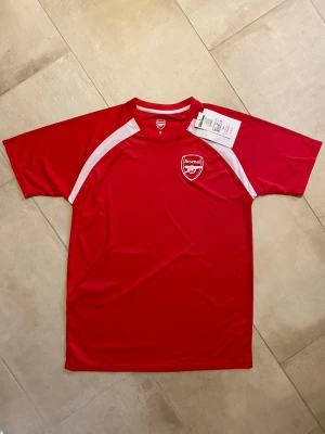 Officiell Arsenaltröja - Hej! Säljer en officiell Arsenaltröja i storlek S. Den är tillverkad i skönt och ventilerande funktionsmaterial. Den är aldrig använd och lappen sitter kvar. Perfekt som budgetalternativ för sommarens fotbollsäventyr! Tveka inte att höra av dig vid frågor!