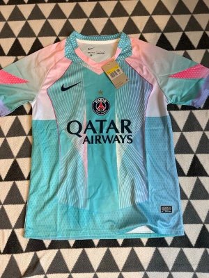 PSG matchtröja i pastell från Nike - Säljer en PSG matchtröja från Nike i snygga pastellfärger med turkos, rosa och lila toner. Tröjan har korta ärmar, Dri-Fit material och Paris Saint-Germain-logga på bröstet. Perfekt för fotboll eller streetwear.