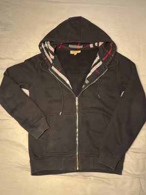 Svart hoodie från Burberry med dragkedja - Svart hoodie från Burberry med klassiskt rutigt foder i huvan och dragkedja framtill. Mjukt material, två fickor och justerbar huva med snören. Perfekt för en stilren och avslappnad look.