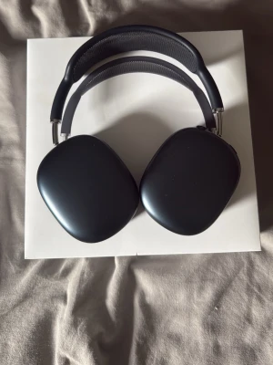 AirPods max - Stilrena svarta over-ear hörlurar med mjuka öronkuddar och justerbart huvudband i metall och textil. Modern design med rundade former och mattsvart finish. Perfekta för musik, gaming eller videosamtal.