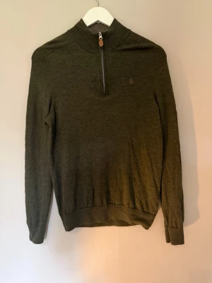 Olivgrön stickad tröja med halv dragkedja - Snygg olivgrön stickad tröja med hög krage och halv dragkedja framtill. Tröjan har långa ärmar, ribbade muddar och en liten broderad logga på bröstet. Perfekt för lager på lager och enkel att matcha med jeans eller chinos.