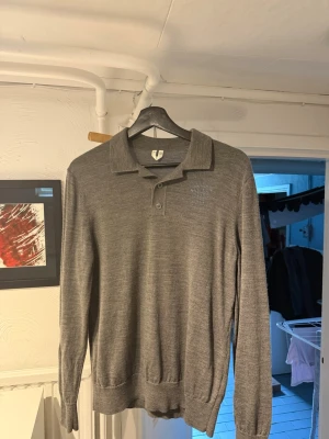Grå långärmad pikétröja från Arket - Stilren grå långärmad pikétröja med krage och knappar framtill. Tröjan är tillverkad i ett tunt, mjukt material och har ribbade muddar vid ärmslut och nederkant. Perfekt för dig som gillar en clean och enkel look.