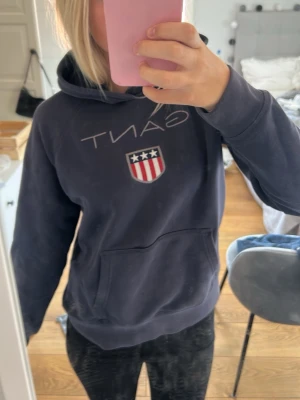 Mörkblå hoodie från GANT med logga - Säljer en mörkblå hoodie från GANT med stor logga och amerikanskinspirerat sköldmärke på bröstet. Hoodien har huva och en stor magficka. Tillverkad i mjukt bomullsmaterial och har ribbade muddar vid ärmslut och nederkant. Perfekt för en avslappnad look.