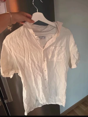 Vit kortärmad linneskjorta - Säljer en vit skjorta i linne med korta ärmar och knappar framtill. Skjortan har en klassisk krage och en bröstficka. Perfekt för varma dagar och enkel att styla till olika outfits.