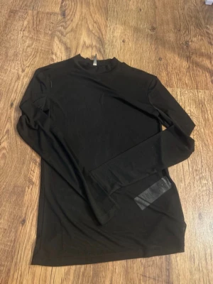 Svart mesh polotopp med lång ärm - Snygg svart polotopp i meshmaterial med lång ärm. Toppen är genomskinlig och har en stilren, tight passform som passar perfekt till lager-på-lager-stil. Enkel och trendig design utan synliga detaljer.