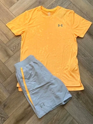Under Armour träningsset gul/grå - Snyggt träningsset från Under Armour med en gul t-shirt och ljusgrå shorts. T-shirten har rund hals och logga på bröstet, shortsen har elastisk midja, sidofickor och gul rand längs sidan. Perfekt för gymmet eller löprundan.