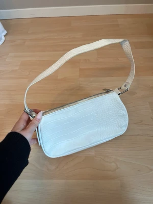 Väska - Snygg vit axelväska i croco-imitation med silverfärgade detaljer. Väskan har en bred axelrem och dragkedja upptill samt en extra ficka med dragkedja på baksidan. Några skavanker som syns på bilderna. 