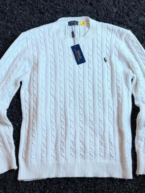 Vit kabelstickad tröja Polo Ralph Lauren - Kabelstickad vit tröja från Polo Ralph Lauren med rund halsringning och klassisk broderad logga på bröstet. Tröjan har ribbade muddar vid ärmslut och nederkant. Perfekt för dig som gillar stilrena och tidlösa plagg.