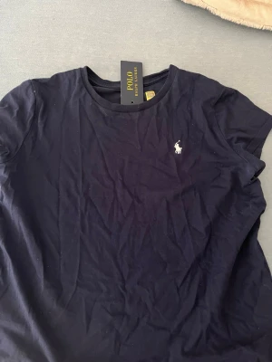 Mörkblå t-shirt från Polo Ralph Lauren - Klassisk mörkblå t-shirt från Polo Ralph Lauren med rund halsringning och ikoniska vita logobroderiet på bröstet. Tillverkad i mjuk bomull och har en normal passform med korta ärmar. Perfekt basplagg både för stilen och sommaren!