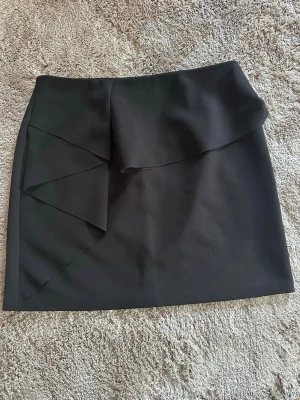 Svart kortkjol med volang från Zara - Säljer en svart kortkjol från Zara med snygg volangdetalj framtill. Kjolen har en enkel och stilren design med en liten slits och stängs med dragkedja bak. Perfekt för dig som gillar trendiga och klassiska plagg.