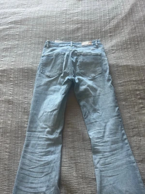 Ljusblå bootcut jeans - Säljer ett par ljusblå bootcut jeans med klassisk femficksdesign och utsvängda ben. Jeansen har en snygg tvätt och är perfekta för dig som gillar retrovibbar. Materialet är denim i bomull. Passar till dig som vill ha en avslappnad men trendig look.