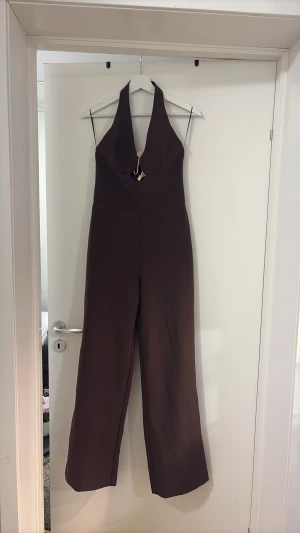 Brun halterneck jumpsuit med cut-out - Snygg brun jumpsuit med halterneck och djup urringning. Framtill finns en cut-out detalj med dekorativt snöre och guldfärgad pärla. Vida ben och figurnära passform upptill. Perfekt för dig som vill sticka ut lite extra.