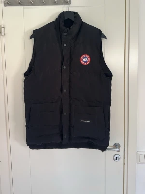 Svart dunväst från Canada Goose sitter som M!! - Snygg svart dunväst från Canada Goose med klassisk logotyp på bröstet och två stora fickor framtill. Västen har tryckknappar och är vadderad för extra värme. Perfekt för lager-på-lager och streetwear-stil. Storlek L sitter som M!!