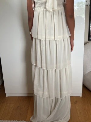 Vit långklänning med volanger - Säljer en elegant vit långklänning med flera lager volanger och halterneck. Klänningen har ett omlottliknande bystparti och är axelbandslös med knyt i nacken. Perfekt för sommarens alla tillfällen!