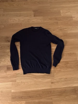 Mörkblå tröja från J.Lindeberg - Klassisk mörkblå tröja från J.Lindeberg med rund halsringning och ribbade muddar vid ärmslut och nederkant. Enkel och stilren design som passar till det mesta. Perfekt basplagg för garderoben. Priset ej hugget i sten!