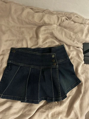 Mörkblå jeanskjol  - Kort jeanskjol i mörkblå denim med tre dekorativa metallknappar framtill. Storlek XS