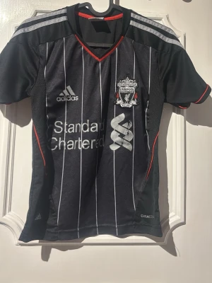Liverpool bortatröja Suarez #7 Adidas - Snygg svart Liverpool bortatröja från Adidas med vita ränder, röda detaljer och tryck av Suarez #7 på ryggen. Tröjan har klubbmärke, sponsorlogga och klassisk V-ringning. Tillverkad i lätt och ventilerande material, perfekt för fotboll eller träning.
