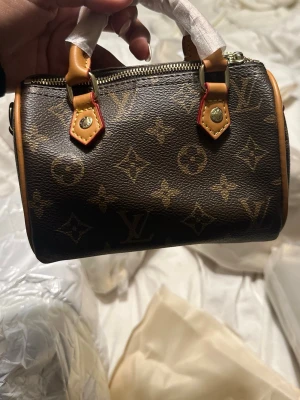 Monogram väska  - Säljer en handväska i klassiskt brunt monogramcanvas med beigea läderhandtag och guldfärgade metalldetaljer. Väskan har cylinderformad design och dragkedja upptill. Perfekt för dig som gillar lyxiga accessoarer och ikonisk stil.