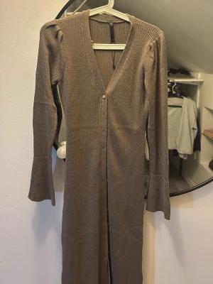 Brun ribbad långklänning med v-ringning - Långärmad brun ribbad långklänning med djup v-ringning och knappar framtill. Klänningen har en figurnära passform och lätt utsvängda ärmslut. Perfekt för dig som gillar stilrena och bekväma plagg.