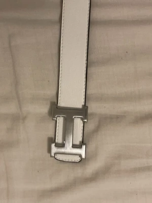 Vitt skärp med silverfärgat H-spänne - Stilrent vitt skärp med ett ikoniskt H-format spänne i silverfärg. Bältet är gjort i skinn och har en klassisk, rak form som passar till många olika outfits. Perfekt accessoar för att lyfta din look.