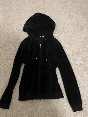 Svart velour hoodie med dragkedja - Snygg svart hoodie i mjuk velour med dragkedja framtill och två fickor. Tröjan har huva med snörning och ribbade muddar vid ärmslut och nederkant. Perfekt för en chill och avslappnad stil.