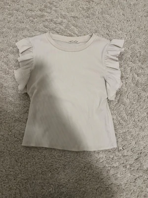 Vit ribbad topp med volangärmar - Supersöt vit ribbad topp med volangdetaljer på axlarna. Toppen har rund halsringning och är ärmlös med breda volanger som ger en trendig look. Perfekt att styla med jeans eller kjol för en fräsch outfit.