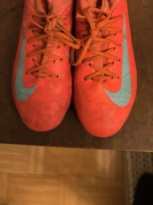 Nike Mercurial röda fotbollsskor - Säljer ett par Nike Mercurial fotbollsskor i rött med stora ljusblå swoosh-loggor på sidorna. Skorna har snörning och låg profil, tillverkade i syntetmaterial för bra grepp och känsla på planen. Perfekta för dig som vill sticka ut på fotbollsplanen och har ett hål på sidan
