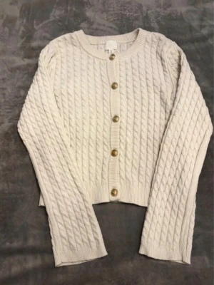 Vit kabelstickad kofta från H&M - Snygg vit kabelstickad kofta från H&M med rund halsringning och guldfärgade knappar framtill. Koft,an har långa ärmar och ribbade muddar vid ärmslut och nederkant. Perfekt att slänga över en t-shirt för en chill look.