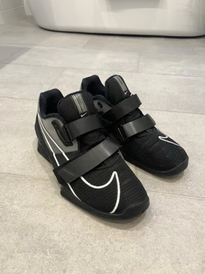 Nike svart träningssko med kardborre - Säljer ett par svarta träningsskor från Nike med vita detaljer och två breda kardborreband över foten. Skorna har en sportig design, meshmaterial för bra ventilation och en stabil sula med unik struktur. Perfekta för gym eller tyngdlyftning. Skorna är i stolek 44 men passar dem som har storlek 43