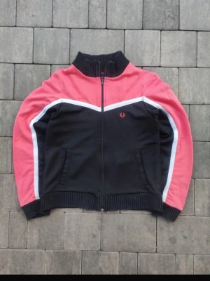 Svart och rosa ziptröja Fred Perry - Snygg ziptröja från Fred Perry i svart och rosa med vita detaljer längs ärmarna. Tröjan har ribbade muddar, hög krage och två fickor framtill. Ikoniska Fred Perry-loggan broderad på bröstet. Perfekt för en sportig och avslappnad stil.