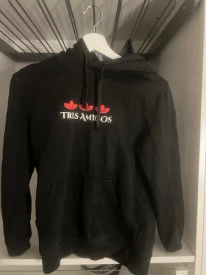 Svart hoodie Tres Amigos - Svart hoodie med huva och magficka från Tres Amigos. Tryck framtill med tre röda sombreros och texten 'TRES AMIGOS' i vitt. Klassisk passform och mjukt material, perfekt för kalla dagar. Det är en limited edition hoodie av 3 personer från paradies hotel.