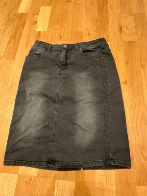 Svart jeanskjol från Flash Woman - Svart jeanskjol från Flash Woman med klassisk femficksdesign och knapp framtill. Kjolen är tillverkad i en mix av bomull, polyester och spandex för en bekväm passform. Enkel och stilren modell som passar till många olika looks.