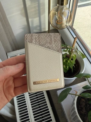 Beige korthållare från iDeal of Sweden - Stilren korthållare i beige och ljusgrått från iDeal of Sweden. Plånboken har en elegant struktur med ormmönstrad detalj upptill och guldfärgad logotyp framtill. Tillverkad i konstläder med plats för flera kort.