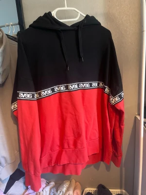Röd och svart hoodie från Svea - Snygg hoodie från Svea i rött och svart med huva och dragsnören. Har en bred logotypremsa med Svea-tryck över bröstet och ärmarna. Perfekt för en avslappnad streetstil. Mjuk bomullskänsla och rymlig passform.