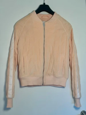  Bomberjacka Lorna Jane - Ljusrosa bomberjacka från Lorna Jane med quiltat tyg, dragkedja framtill och ribbade muddar. Jackan har vita ränder längs ärmarna och två fickor fram. Tillverkad i mjukt material för en sportig och trendig stil.