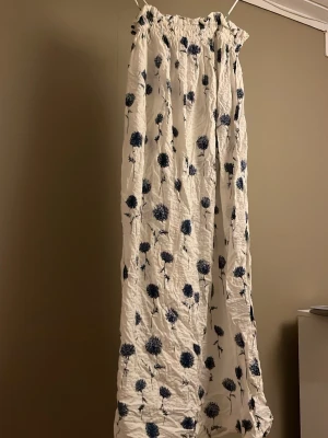 Vit långklänning med blå blommor - Långklänning i vitt med blått blommönster och smock upptill. Klänningen har axelband och ett luftigt, rakt fall som ger en avslappnad känsla. Perfekt för varma dagar och somriga tillfällen.