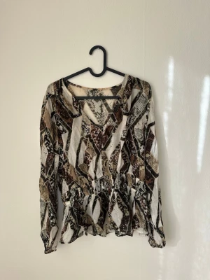 Mönstrad blus - Säljer en snygg blus i beige, brunt och svart. Blusen har v-ringning, resår i midjan och långa ärmar. Materialet är tunt och lätt