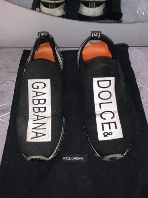 Dolce & Gabbana slip-on sneakers - Svarta slip-on sneakers från Dolce & Gabbana med stora vita logotexter på ovansidan. Skorna har en sportig design med meshmaterial, elastisk öppning och orange innersula. Sulan är svartvit med coola detaljer på sidorna.